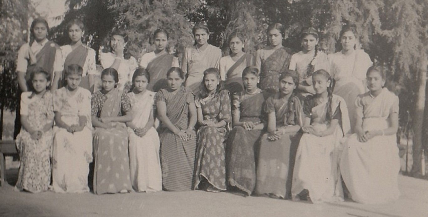 A historical black and white photograph of a group of young women and men posed together outdoors, dressed in traditional attire, reflecting a sense of community and cultural heritage.