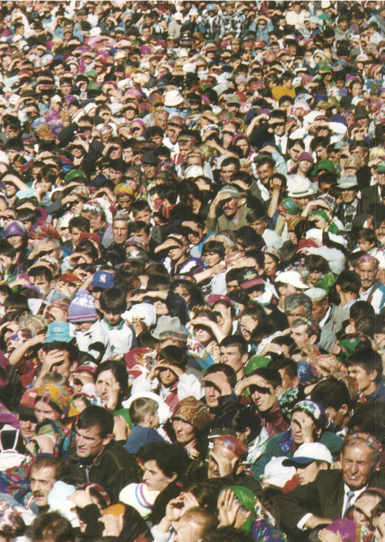 A large crowd of people, many wearing hats and colorful headscarves, gathered outdoors, shielding their eyes from the sun.