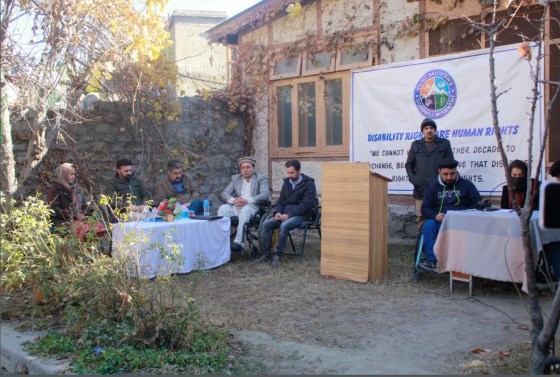 A group of individuals seated in a garden area, with a banner in the background emphasizing disability rights and human rights. The setting includes chairs and a podium, surrounded by foliage, suggesting a community meeting or discussion.