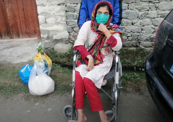 A woman in a wheelchair wearing a protective mask sits outdoors with bags of supplies nearby, showcasing the efforts for support and inclusion for disabled individuals.