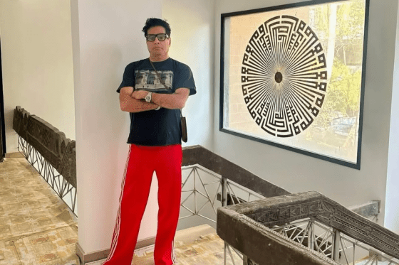 A man standing in a stylish interior space, wearing a black T-shirt and red track pants. The background features a decorative wall with a circular artistic design and a railing that resembles modern architecture.