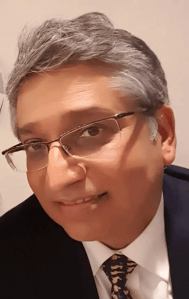 A portrait of Alnoor Jehangir Merchant, a librarian and independent researcher, smiling while wearing glasses and a formal suit.