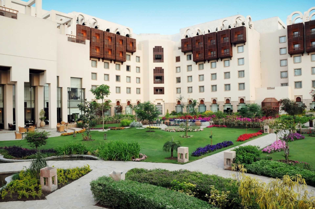 A picturesque courtyard of a hotel featuring well-maintained gardens with colorful flowers, lush green lawns, and modern architecture in the background.
