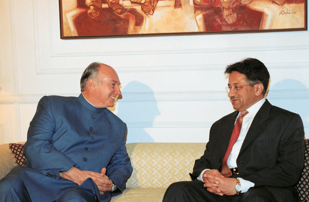 Two men engaged in a conversation while seated on a sofa, with an abstract painting visible in the background.