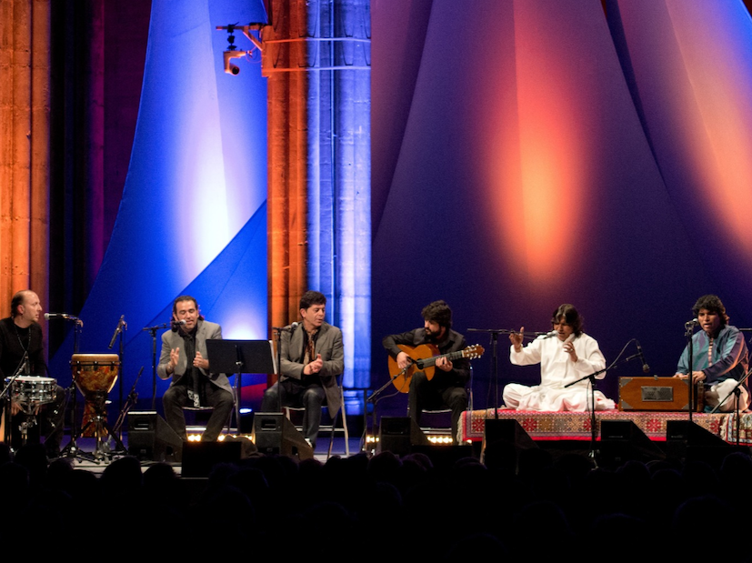A group of five musicians performing on stage, surrounded by colorful lighting. One musician plays a percussion instrument, while another strums a guitar. The central performer is seated on a platform with a harmonium, and the scene captures a live music performance atmosphere.